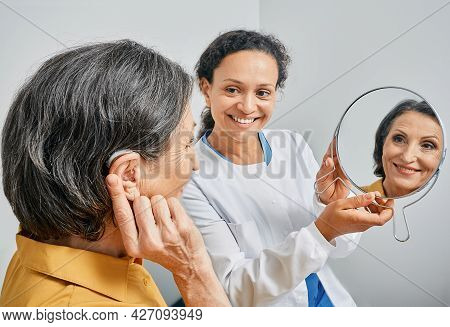 Mature Woman With Bte Hearing Aid Looks At Himself In Mirror Held By Smiling Audiologist, And Tries 