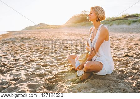 Blonde Woman Wearing Long White Dress Sit At Empty Beach And Look At The Sea
