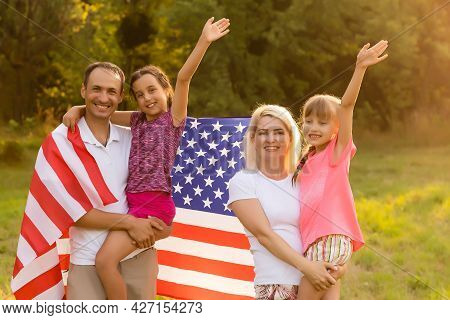 Happy Family Sitting Together In Their Backyard Holding The American Flag Behind Them. Smiling Coupl