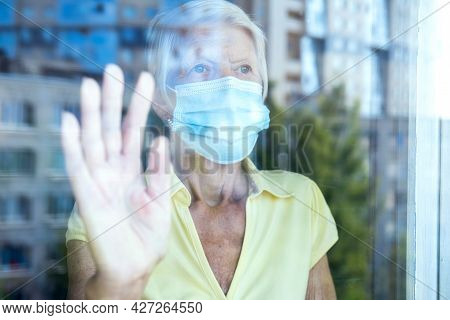 Elderly Caucasian Women Wearing Protective Face Mask, In Home, Looking Outside Window. Old Lady Sadn