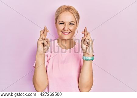Young blonde woman wearing casual pink t shirt gesturing finger crossed smiling with hope and eyes closed. luck and superstitious concept. 