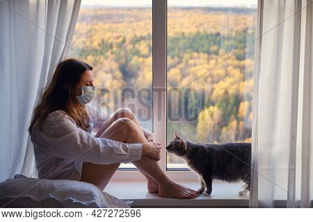 Adult Woman Sits On A Window Sill In A Face Mask In Quarantine And Cat. Sad Autumn Weather And Home 