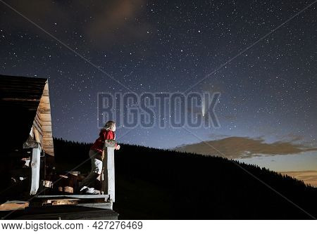 Young Woman In The Mountains Enjoying Beautiful Starry Night And A Comet From A Wooden Hut Porch. Co