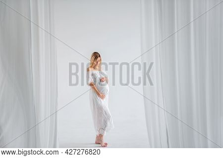 A Young Pregnant Woman In Anticipation Of A Baby In A White Dress, Hugs Belly. Fashion Girl In A Min