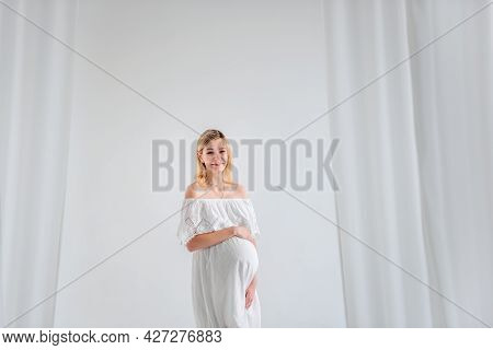 A Young Pregnant Woman In Anticipation Of A Baby In A White Dress, Hugs Belly. Fashion Girl In A Min