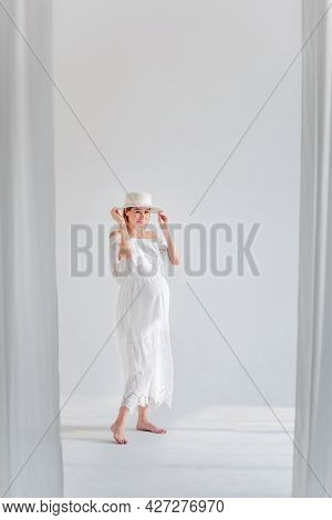Young Pregnant Woman In Anticipation Of A Baby In A White Hat, Dress, Hugs Her Belly. Tender Blonde