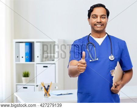 healthcare, profession and medicine concept - happy smiling indian doctor or male nurse in blue uniform with clipboard and stethoscope showing thumbs up over medical office at hospital background