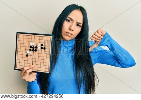 Young hispanic girl holding asian go game board with angry face, negative sign showing dislike with thumbs down, rejection concept 