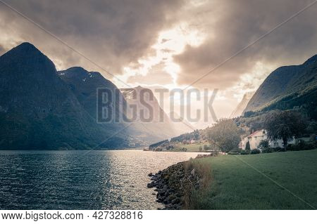 Stormclouds With Sun Shining Through Them Above Dramatic Mountain Range Of Norwegian Fjord. Viewed F