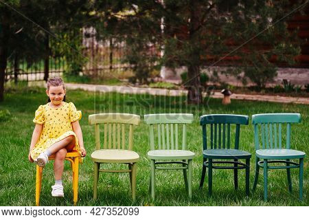 A Funny Girl In A Yellow Dress Is Sitting On A Row Of Chairs Standing On The Lawn In The Park And Pr