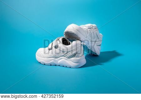 Side View Of A Balanced Composition Of Two White Kid Sneakers With Velcro On A Blue Paper Background
