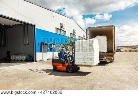 A Man On A Forklift Works In A Large Warehouse, Unloads Bags Of Raw Materials Into A Truck For Trans