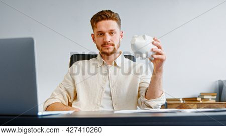 Serious man corporate company manager with beard holds white piggy bank and smiles joyfully posing at office table with laptop