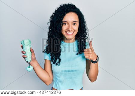 Young hispanic woman with curly hair holding closed umbrella smiling happy and positive, thumb up doing excellent and approval sign 