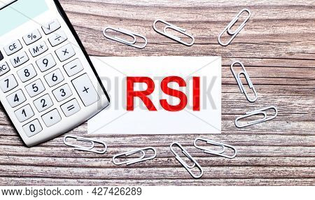 On A Wooden Background, A White Calculator, White Paper Clips And A White Card With The Text Rsi Rel