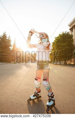 Vertical Low Angle Shot Of A Girl In Protective Gear And Rollerblades, Drinking Water On Hot Summer 