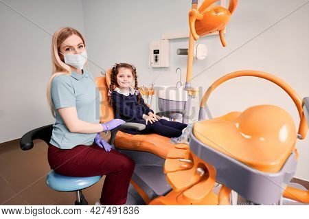 Cute Female Kid Sitting In Dental Chair Beside Stomatologist. Adorable Little Girl Having Appointmen