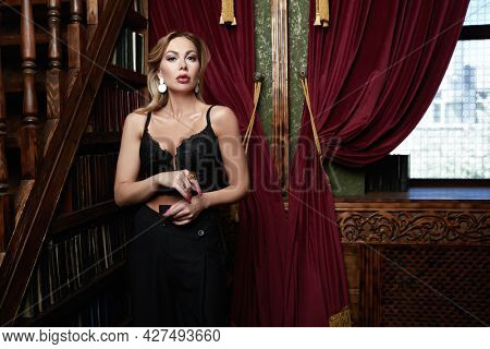 Wealthy middle-aged woman with evening makeup and hairstyle stands in black dress in a luxury apartment. Luxury lifestyle. 