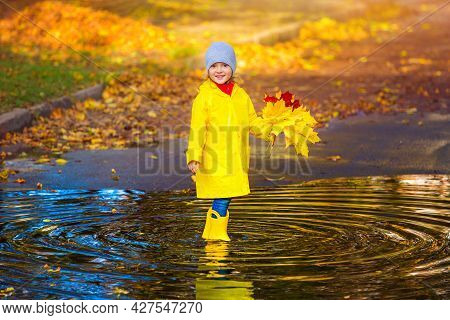 A Happy Girl Child In A Yellow Raincoat And Rubber Boots In A Puddle On An Autumn Walk. Kid With Aut