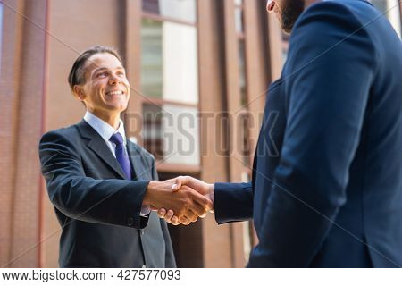 Confident businessman and his colleague are shaking hands in front of modern office building. Financial investors outdoor. Banking and business.