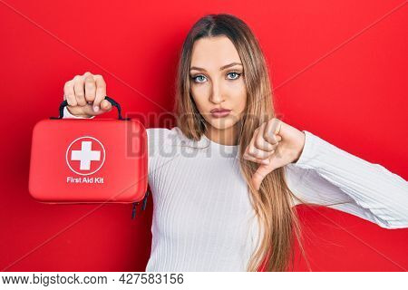 Young blonde girl holding first aid kit with angry face, negative sign showing dislike with thumbs down, rejection concept 