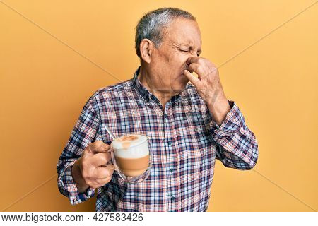 Handsome senior man with grey hair drinking a cup coffee smelling something stinky and disgusting, intolerable smell, holding breath with fingers on nose. bad smell 