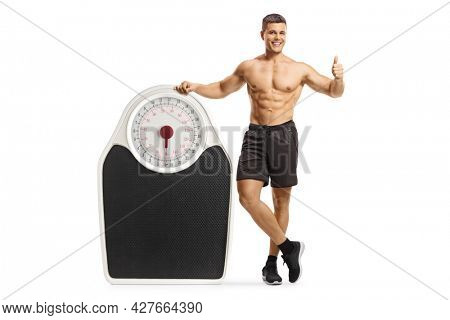 Shirtless young man leaning on a big weigth scale and gesturing thumbs up isolated on white background 