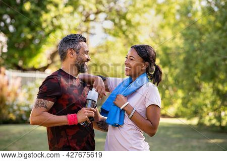Cheerful multiethnic couple resting together after jogging in the park. Happy mature man and beautiful woman laughing while resting after running. Woman and indian man taking a break after fitness.