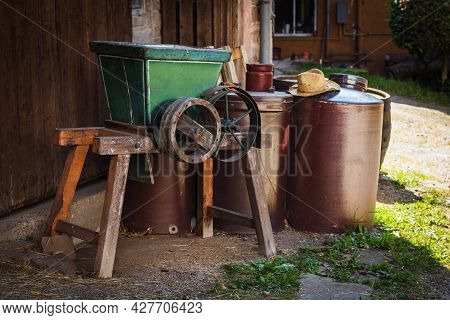 Agricultural Objects In The Yard Of An Old Farmhouse With Grain Crasher And Barrels