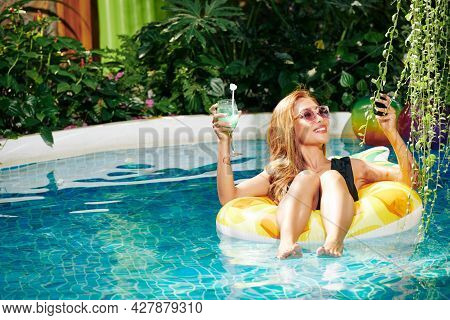 Smiling Attractive Young Woman Lying On Infalible Toy And Taking Selfie With Cocktail