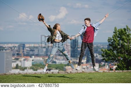 Cheerful Young Couple Travelers In City On Holiday, Jumping. Cityscape In The Background.