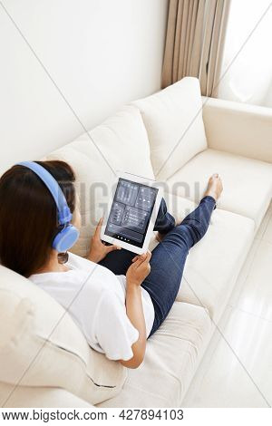 Young Woman In Headphones Sitting On Sofa And Using Application On Tablet Computer To Set Music Volu