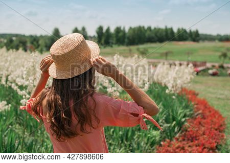 Woman In Hat Near Flower Bed. Girl In Pink Dress, In Straw Hat Stands With Her Back Facing The Sun I
