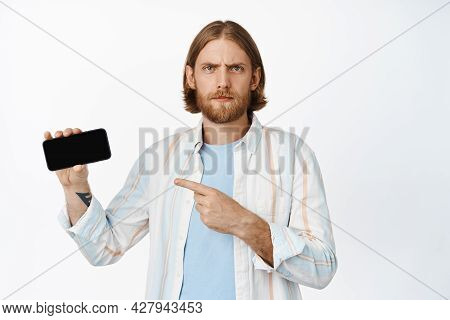 Image Of Confused, Blond Guy Frowning, Pointing At Mobile Phone, Horizontal Screen With Concerned Fa