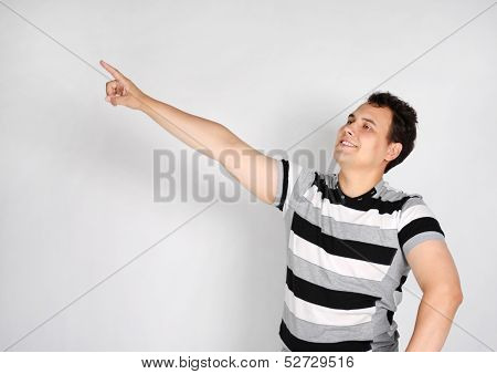 Brunet Man In Striped T-shirt Smiles And Points To Side On Grey Background.