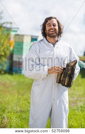 Portrait of happy male beekeeper holding smoker while standing at apiary