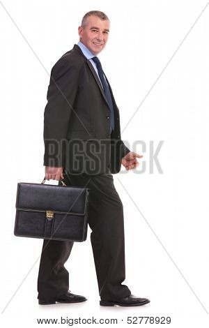 full length side view picture of a business man walking with a briefcase in his hand and looking at the camera with a smile on his face. on a white background