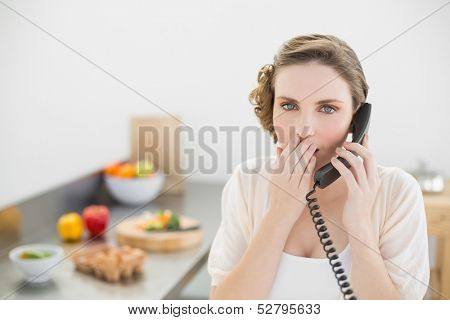 Shocked cute woman phoning in her kitchen with a telephone looking at camera