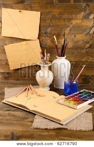 Composition with colorful watercolors, brushes in vase and sketcher on wooden background 