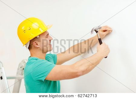 reapir, building and home renovation concept - smiling man in yellow protective helmet hammering nail in wall