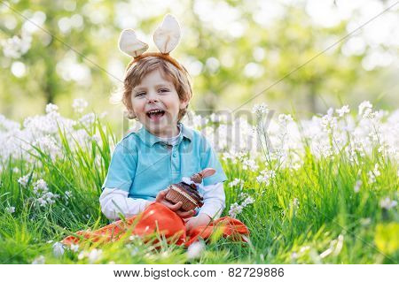 Cute Happy Little Boy Wearing Easter Bunny Ears And Eating Chocolate At Spring Green Grass