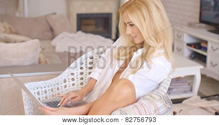 Pretty sexy blond woman relaxing in a white wicker chair in her living room working on a laptop computer with a smile  side view