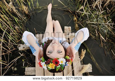 Girl In A Flower Wreath On His Head Sitting On The Bridge And Wets Feet In The River