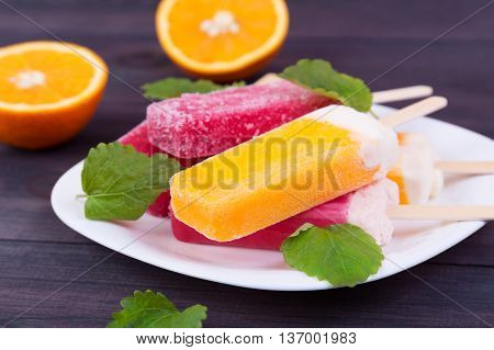 Colored popsicles on a plate with mint and oranges on the wooden background
