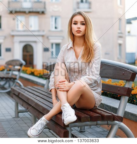 Petty Woman In White Sneakers Sits On A Bench On The Background Of The Building.
