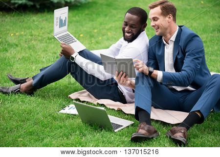 In a good mood. Cheerful smiling overjoyed colleagues sitting on the grass and using laptops while discussing project