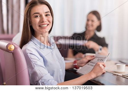 In pace with time. Cheerful charming delighted woman sitting at the table and holding newspaper while her colleague working in the background