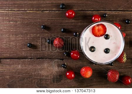 glass of berry yogurt smoothie with fresh ripe berries around on wooden table. top view