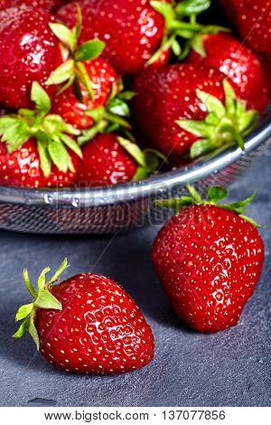 Washed strawberries in sieve with two fresh and juicy strawberries near it on stone background.