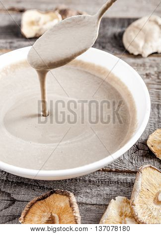 Creamy vegan mushroom soup with champignon and shiitake in white bowl. Soup drains from spoon into bowl. Wood background. Dry shiitake on front.